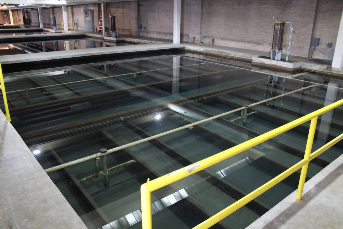 Large indoor water treatment facility with rectangular concrete tanks filled with clear water, surrounded by yellow safety railings and industrial equipment.