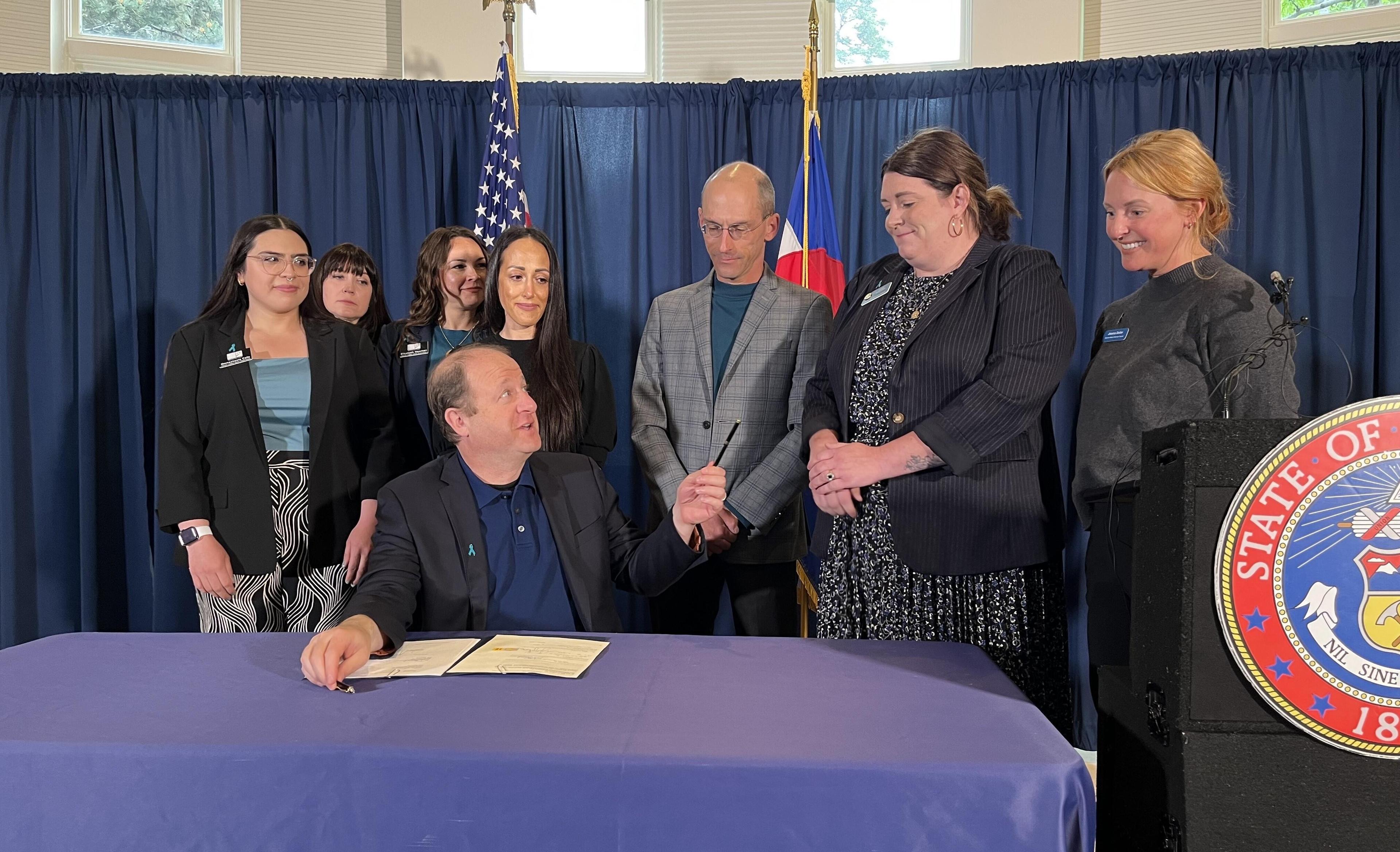 Gov. Jared Polis is seated at a table wearing a dark grey suit and blue shirt. He is looking up at Rep. Jenny Willford, second from right, who is wearing a dark jacket and black and white patterned dress. Sen. Mike Weissman stands behind Polis wearing a light grey jacket. Other people stand around them wearing dark clothes and they are all against a dark blue backdrop.