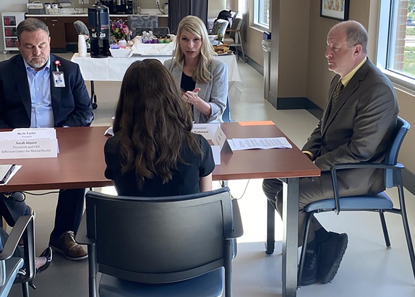 Four people sit around a table. One man in a blue suit jacket and one woman is wearing a beige blazer and making a hand gesture to the woman sitting across from her. A man in a gray suit sit at the head of the table.
