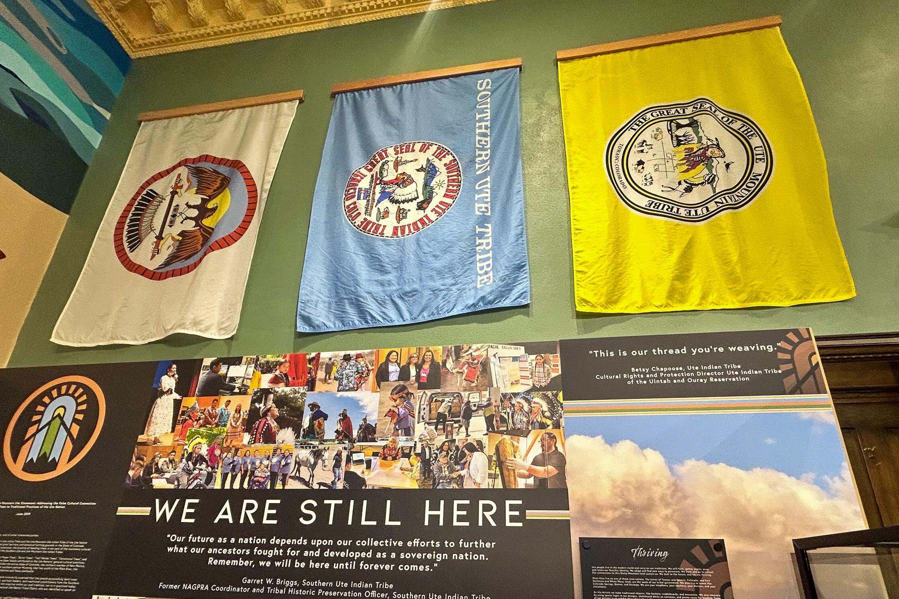 Photo shows three Ute flags hanging above a sign that reads "We are still here" at the Pioneers Museum in Colorado Springs elevating Ute culture and history.
