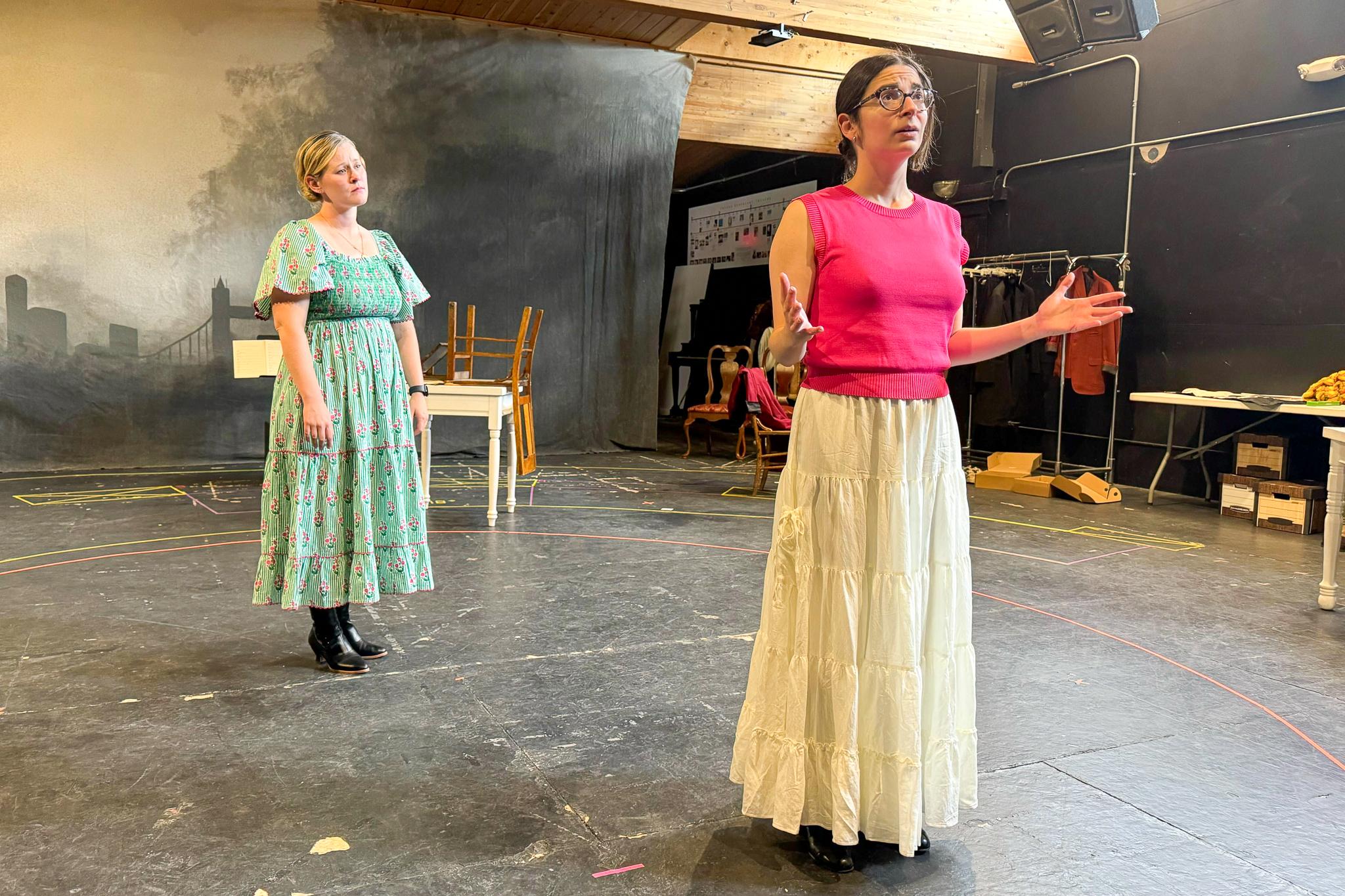 Photo shows 2 actresses on stage. The actress on the right is gesturing and wearing a bright pink top and neutral skirt. The actress on the left in the background is wearing a light green dress.