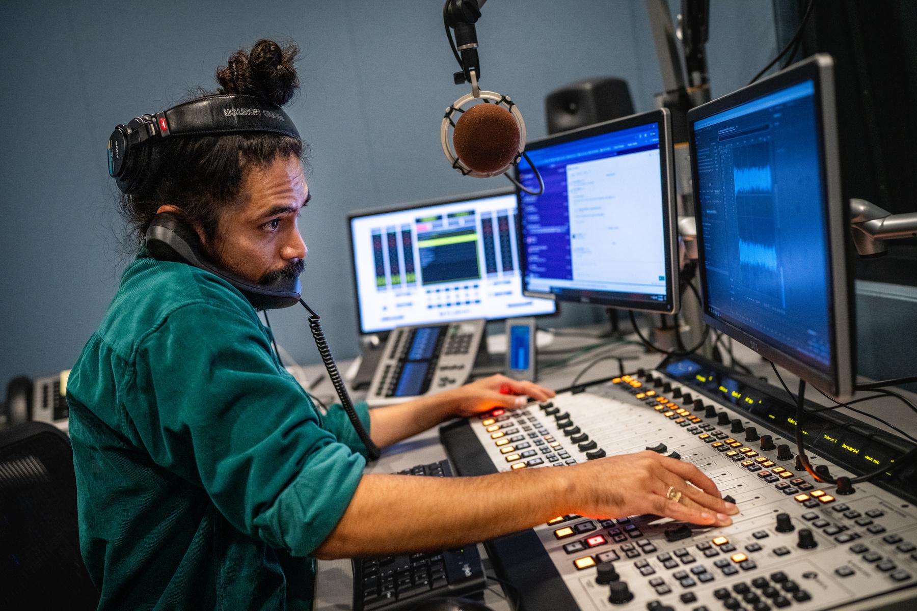 A man in a teal shirt sits at a computer and sound board with a phone up to his ear