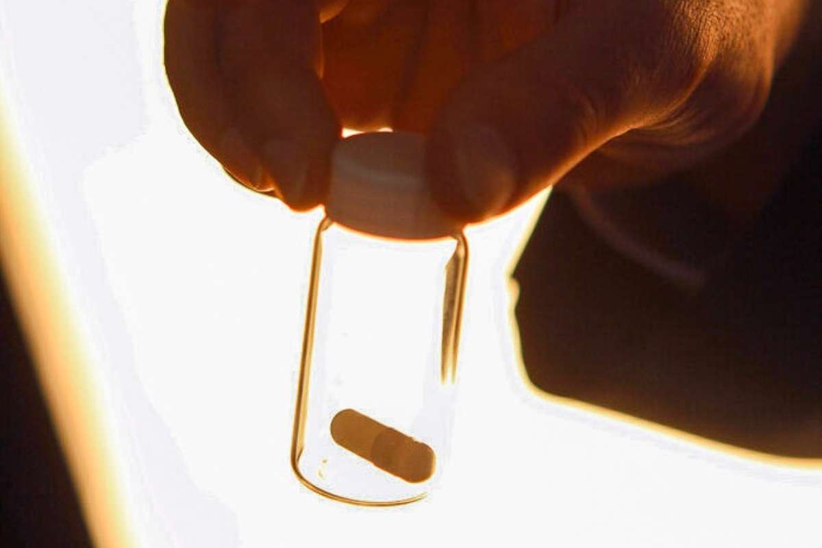 A hand holds a white pill in a clear small bottle with a white lid