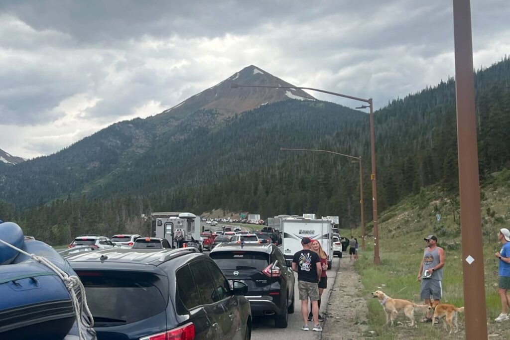 Multiple vehicles stopped along a road with a mountain visible in the background. People are standing outside of their cars