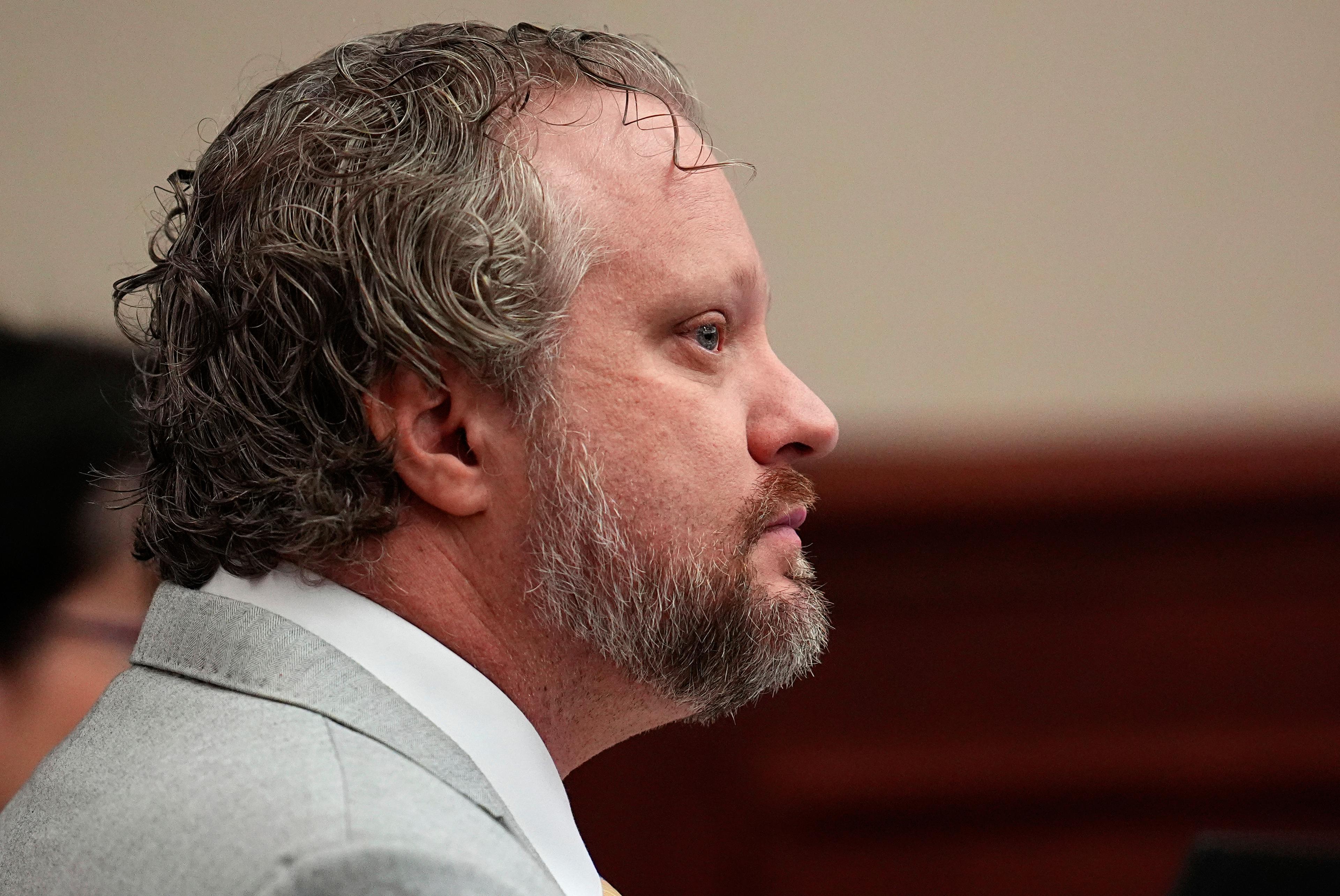 A man sits in a courtroom in a close shot. He wears a gray suit.