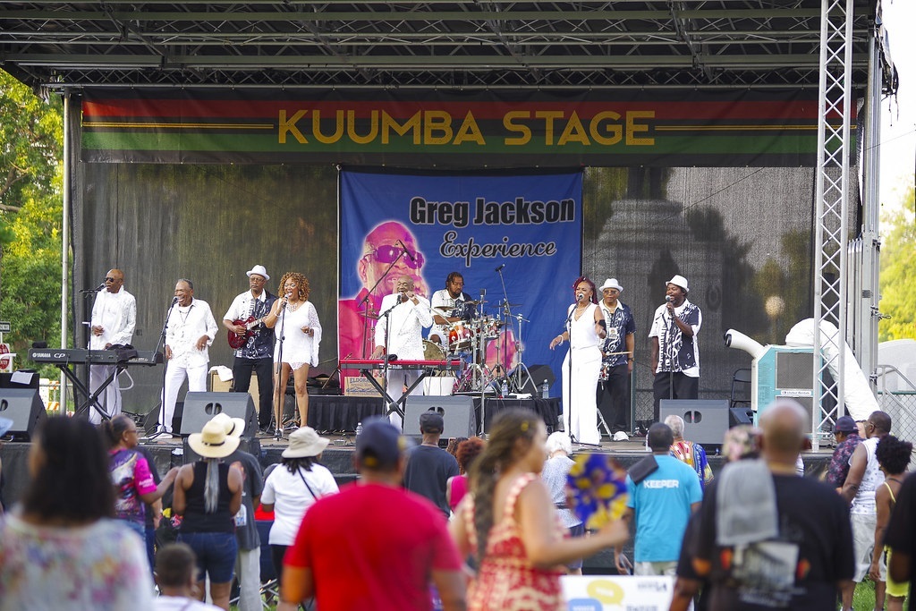 Photo shows a band performing on a stage. The performers are mostly wearing white outfits. Across the top of the stage is a striped banner that reads "Kuumba Stage." There is an audience standing in the foreground.