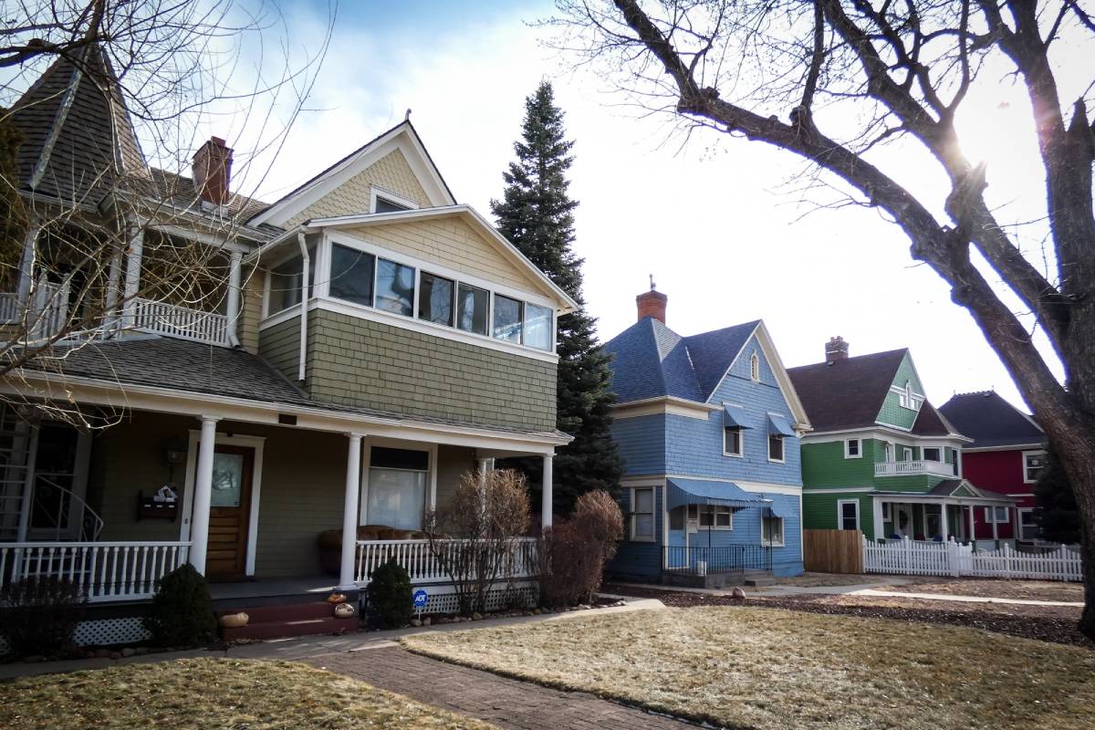 A row of colorful, two-story houses with porches and gabled roofs line a quiet suburban street. Trees without leaves stand in the front yards, and the ground is covered with dry grass