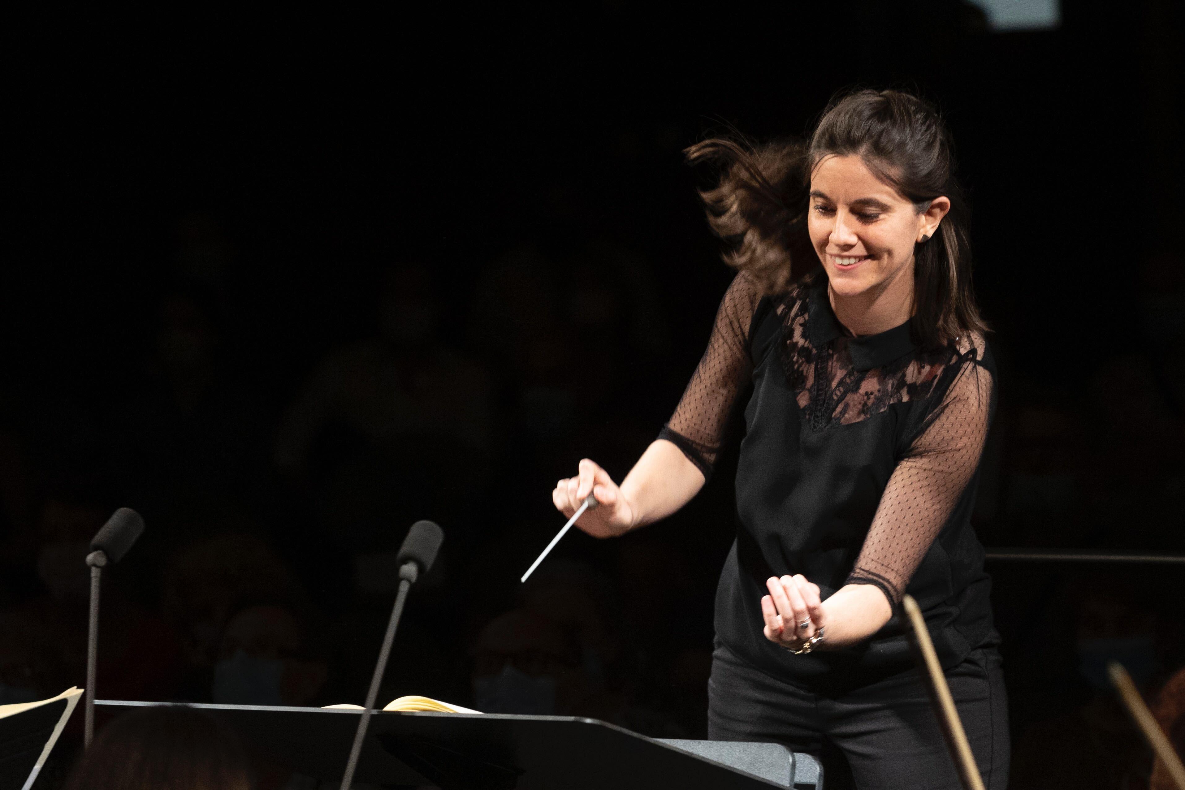 Photo shows a woman with long dark hair wearing a black top with mesh sleeves and black pants in front of a black background directing an orchestra with a conductor's baton in her hand. A microphone is in the front left foreground of the photo.