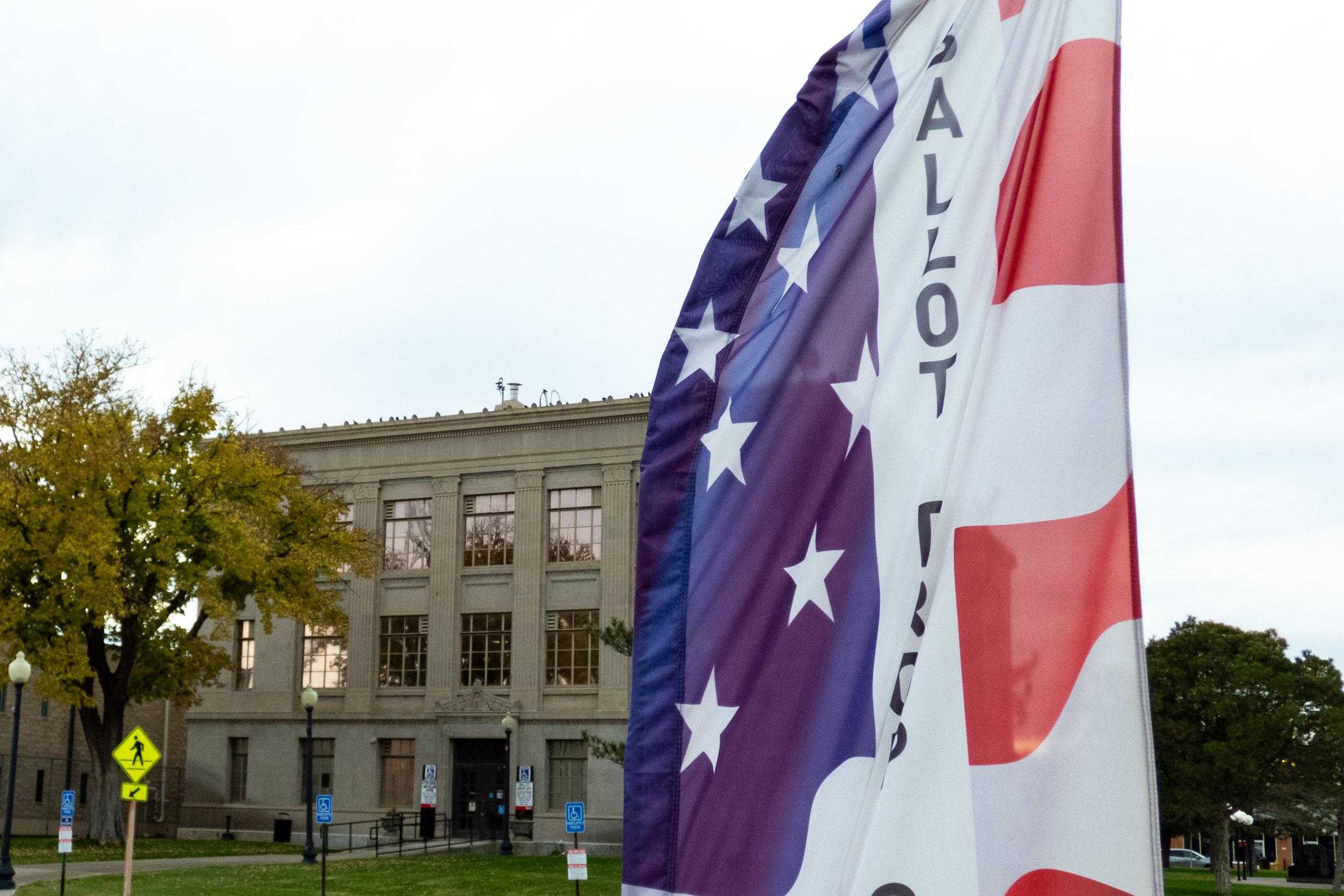 A red white and blue upright banner for balloting in front of a large gray two story building surrounded by trees and grass.