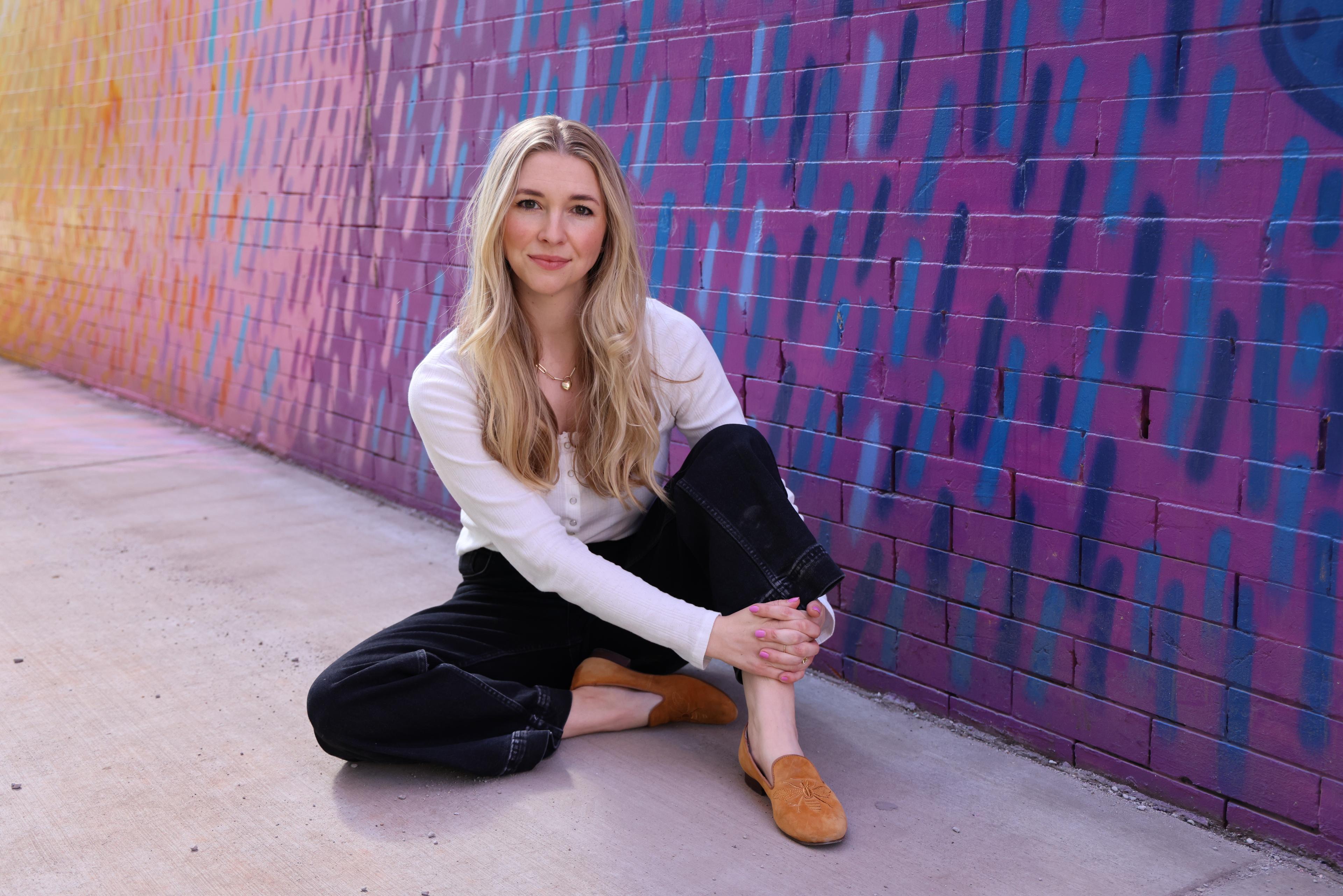 Photo shows a woman with long blonde hair wearing a long sleeve white shirt and black pants sitting on a cement floor with a purple wall angled behind here. There are dark and light blue short diagonal stripes on the wall.