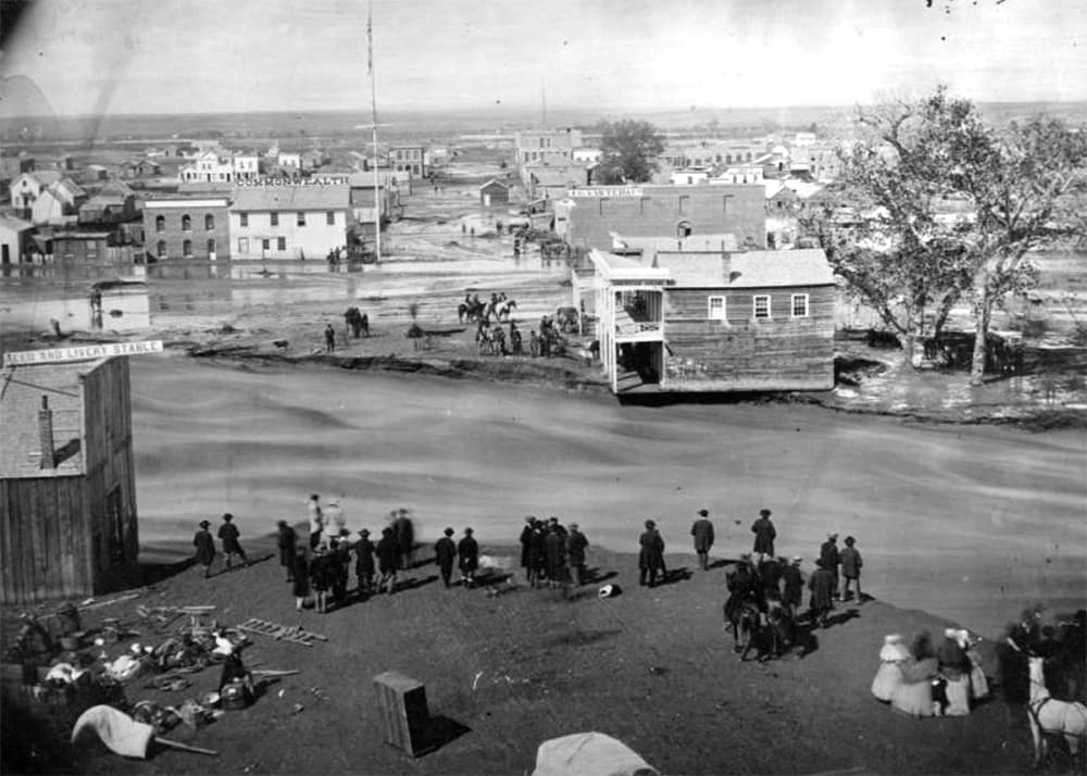 Historic black and white photo shows people in the foreground looking at a flooded creek with a home askew on the shore.