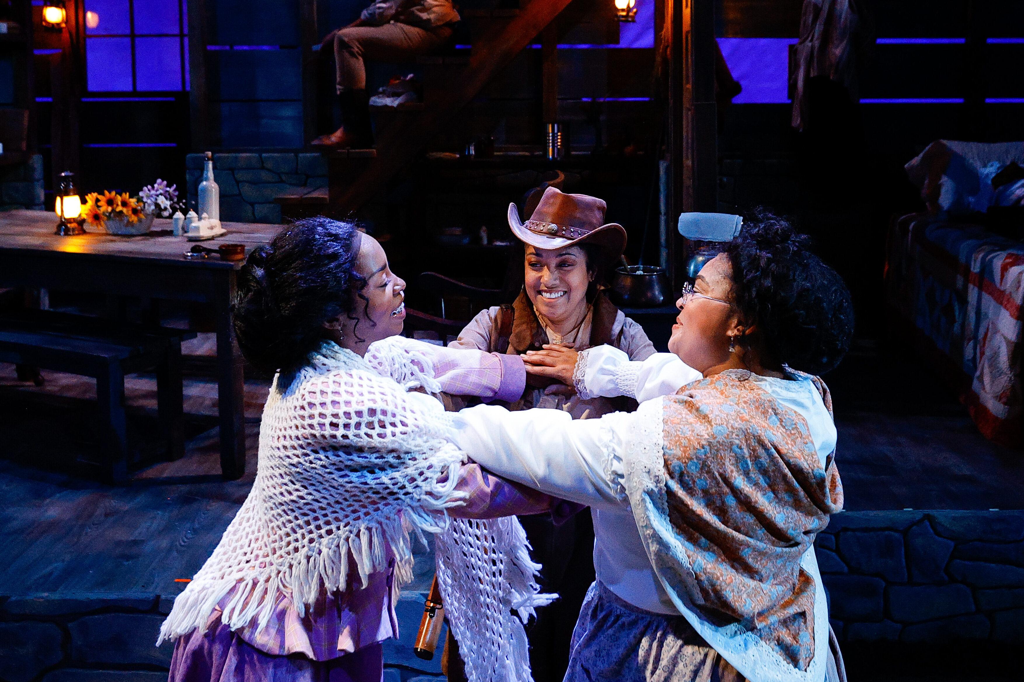 Three actors in 1890s-style clothing stand in a circle, smiling and holding hands on a rustic stage set. Kristina Fountaine as Sophie, Tresha Farris as Minnie, and Cheerish Martin as Fannie perform a scene from Flyin’ West at Theatreworks.