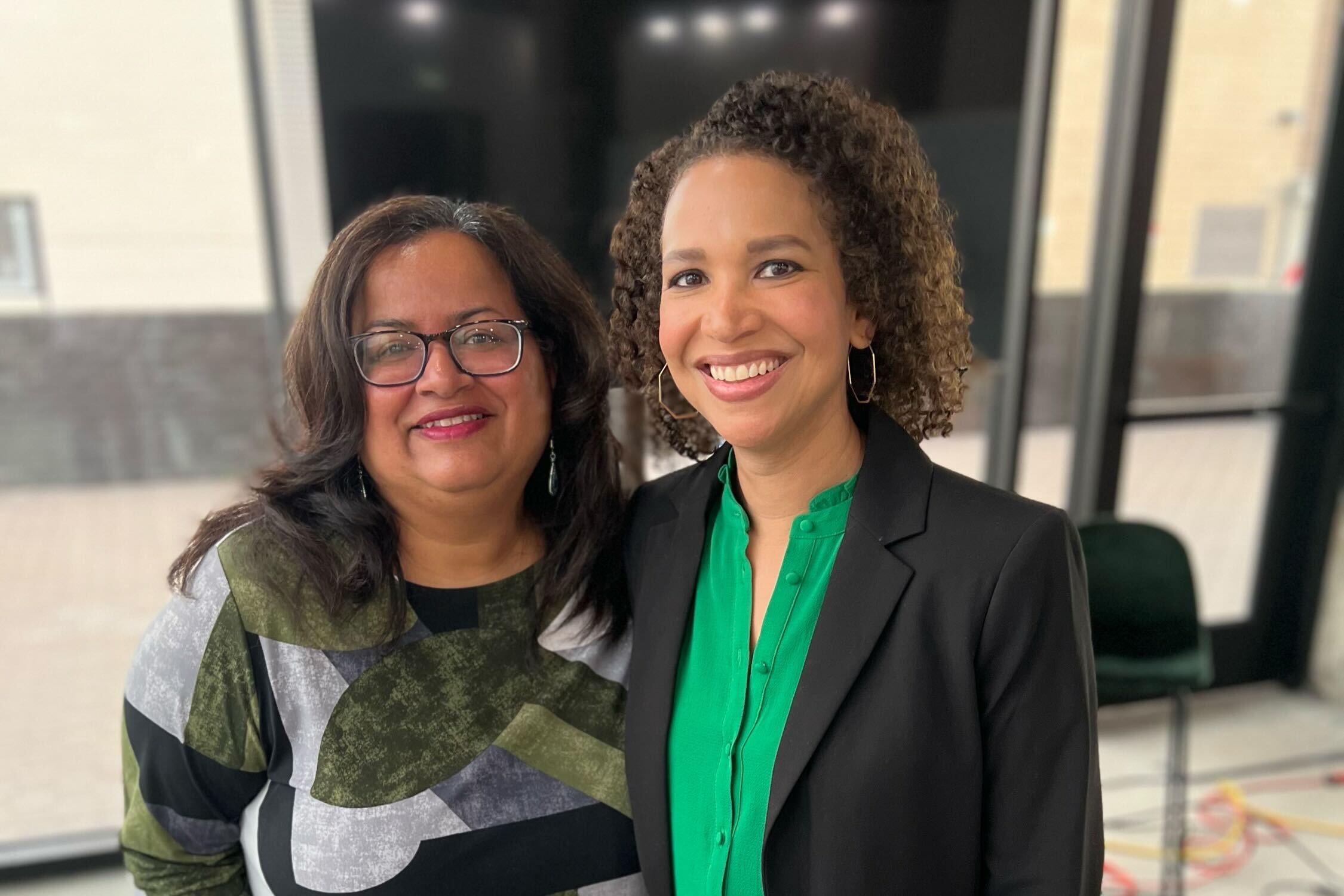 Two women stand in front of a TV screen, with their arms on each other's shoulders. The woman on the right wears a green shirt and black jacket. The woman on the left wears a patterned sweater.