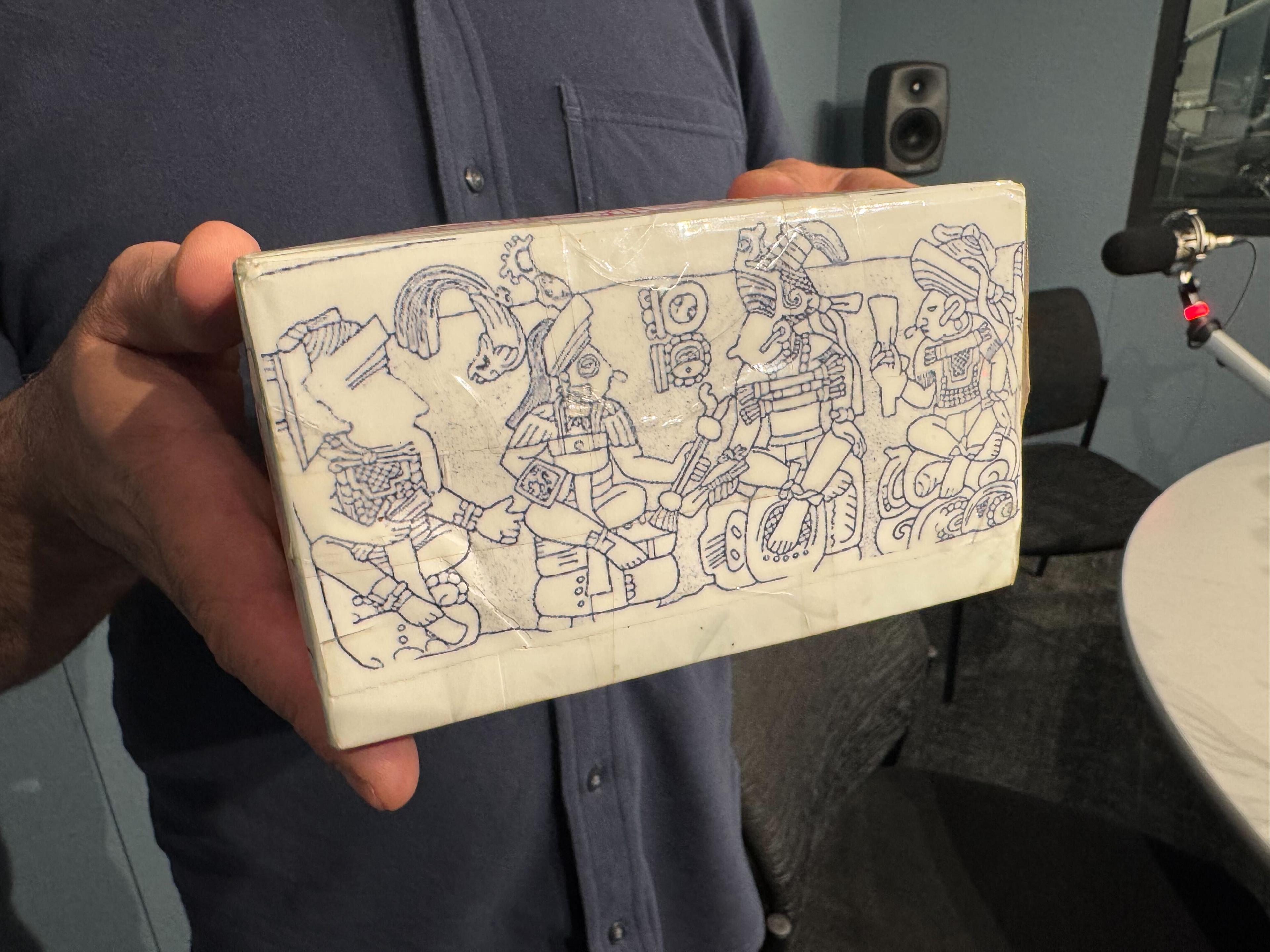 Photo shows hands holding a drawing of stone etchings of Maya sign language. The man holding the sketch is wearing a dark grey shirt in the background.