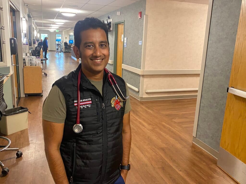 An Indian doctor stands in a hospital hallway. He is smiling and looking at the camera.