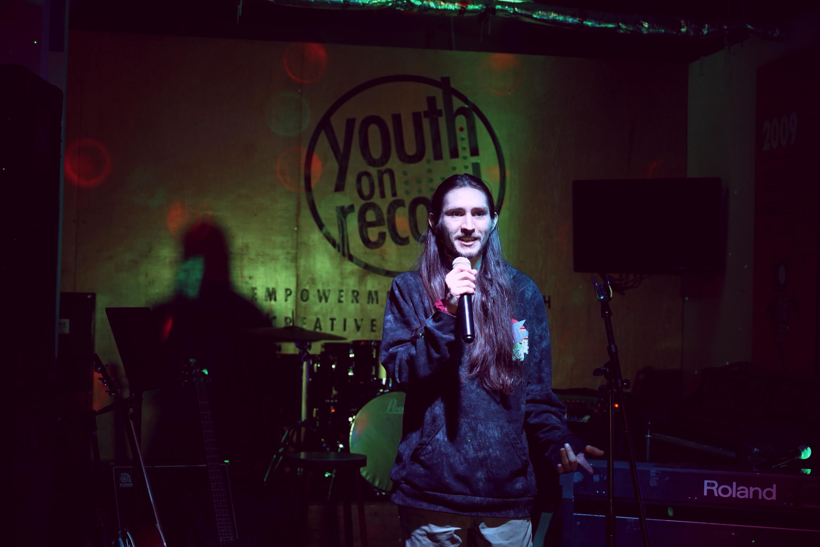 Photo shows a young man in dark clothing standing on a dark stage holding a microphone. The "Youth on Record" logo is behind him.