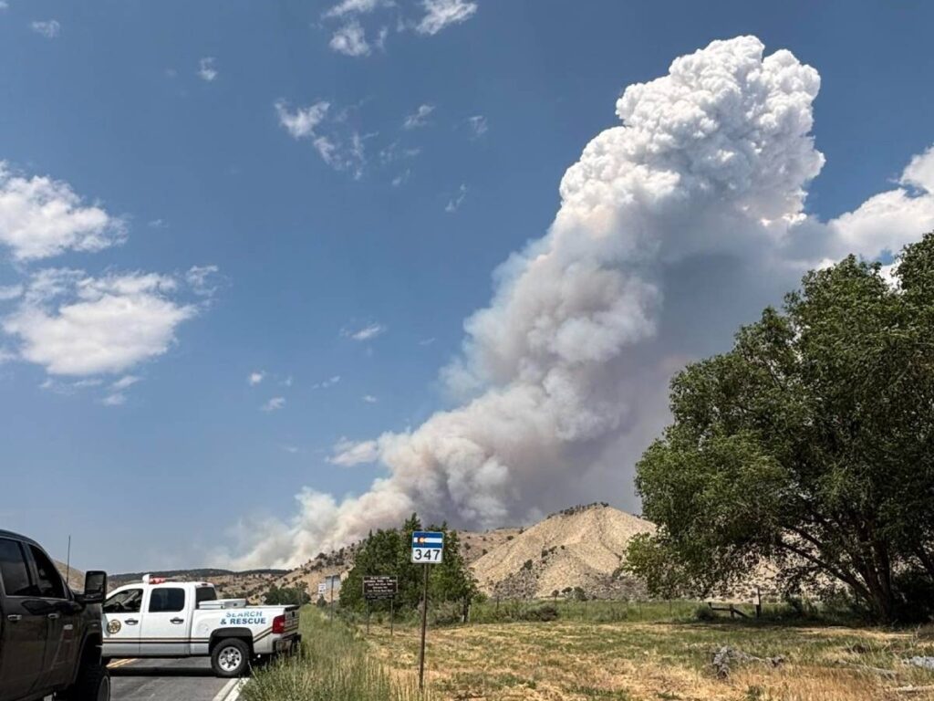 A truck is on a road, with a large plume of smoke rising from the ground, indicating a fire. The sky is clear with a few clouds.