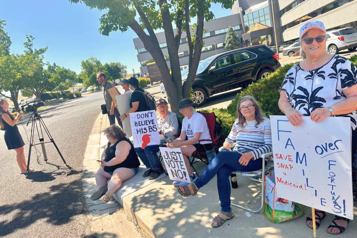 A group of people are sitting on a sidewalk, holding signs and protesting. They are gathered around a tree, with some sitting on chairs and others standing.