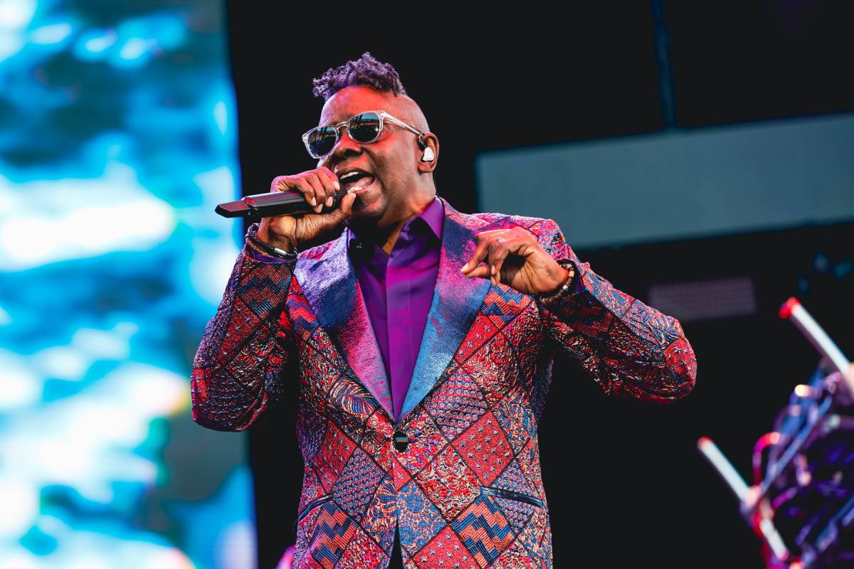 A man with sunglasses in a purple and red plaid suit coat with a purple shirt sings into a microphone on stage
