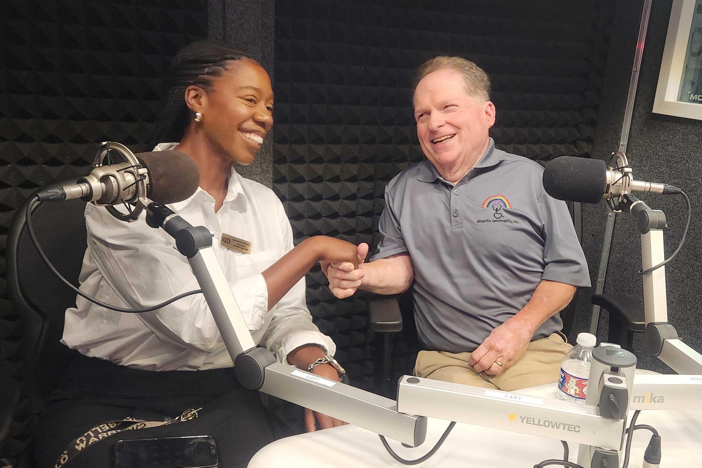 A woman is seated behind a studio microphone and holding hands with a man also seated behind a studio microphone. She is wearing a white shirt and he is wearing a grey shirt. They are looking at each other and smiling.