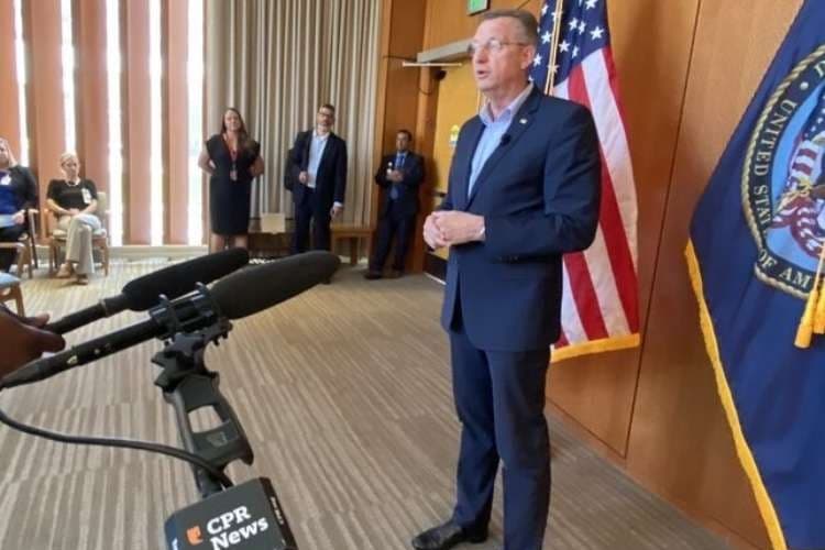 A man in a navy blue suit wearing round wire glasses speaks to a room. Thera are people standing in the background as the man speaks in from on an American flag. In the foreground is a radio microphone.