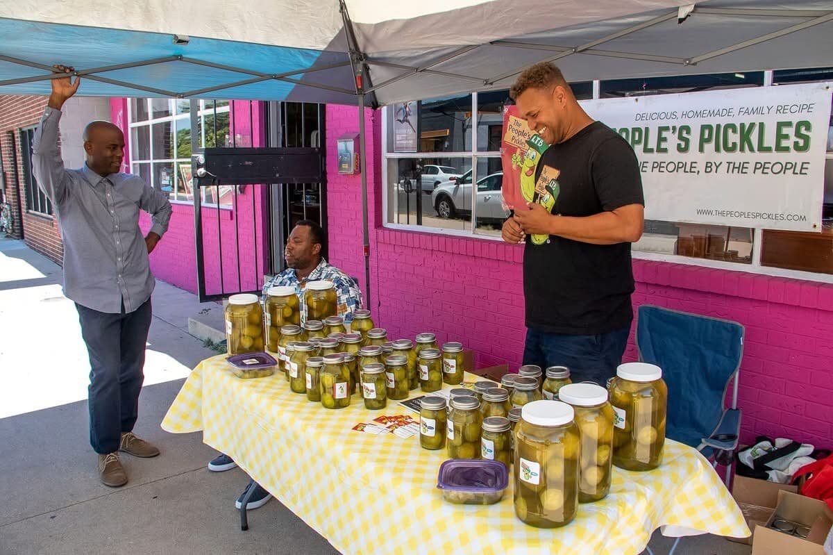 Photo shows a vendor booth outside with a table filled with jars of pickles and am an in black clothing standing to the right.