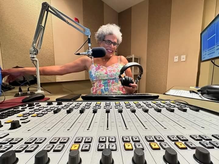 Dana Meyers in a radio broadcast studio, smiling behind a microphone on a boom, with headphones in her hand and a vast control board in front of her.