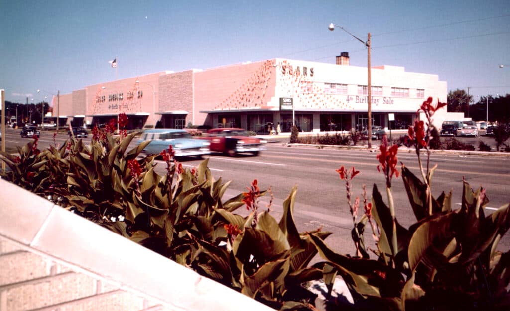 Historic photo has plants in the foreground and the original Sears store in Cherry Creek in the background with a road with cars in front of the store.