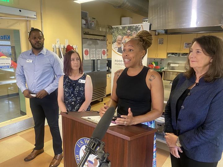 A Black woman stands at a lecturn with the U.S. House of Representatives seaon on it. To the woman's left is a white woman in a navy blue suit; on her tight is a white woman in a dark blue tank top and a Black man in a light blue button-down shirt. All for people are standing in a commercial kitchen.