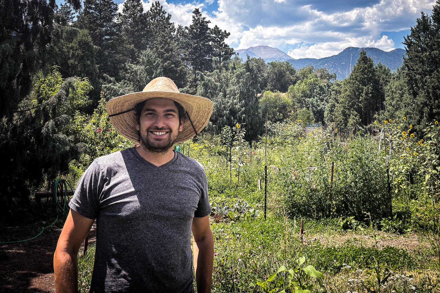 a man in a straw hat stands in front of a garden with trees in the background