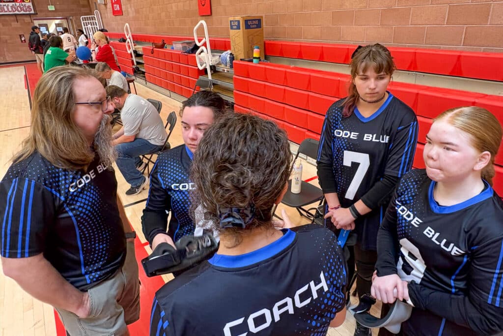 A team of goalball players stands in a circle on the court sidelines.