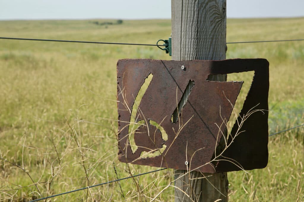 A rusted sign reading “6/7” is nailed to a fencepost.