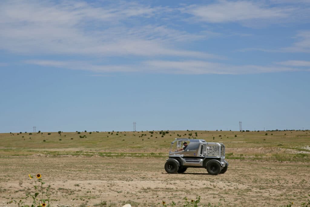 A shining metal moon rover prototype drives across a dusty field.