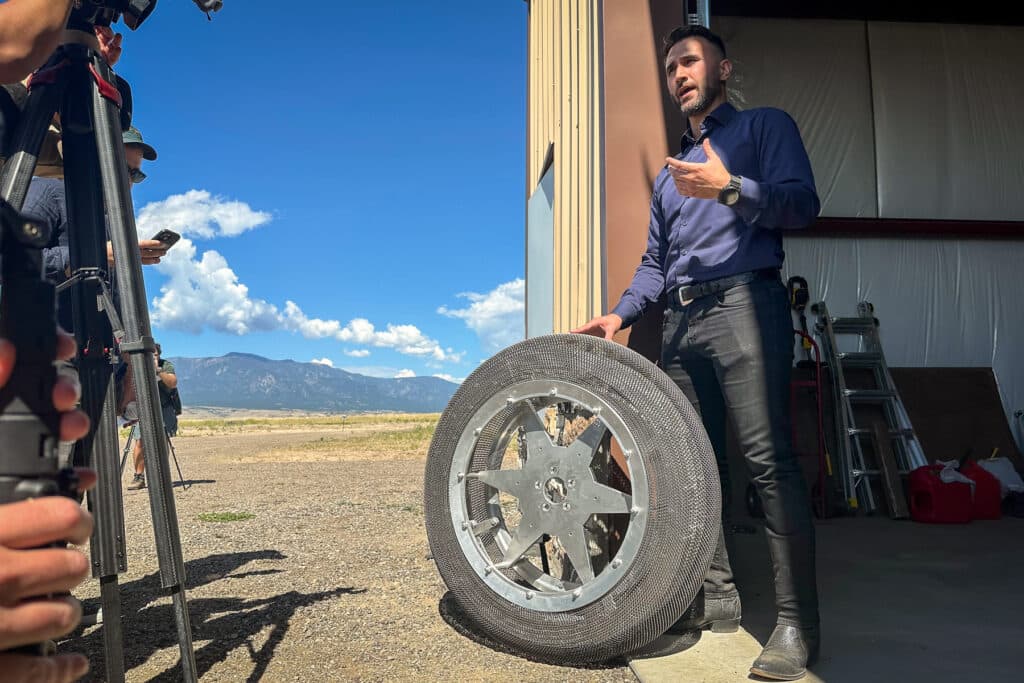 A man in a purple shirt and black pants stands outside a garage next to a tire that uses what appears to be chain-mail instead of rubber.