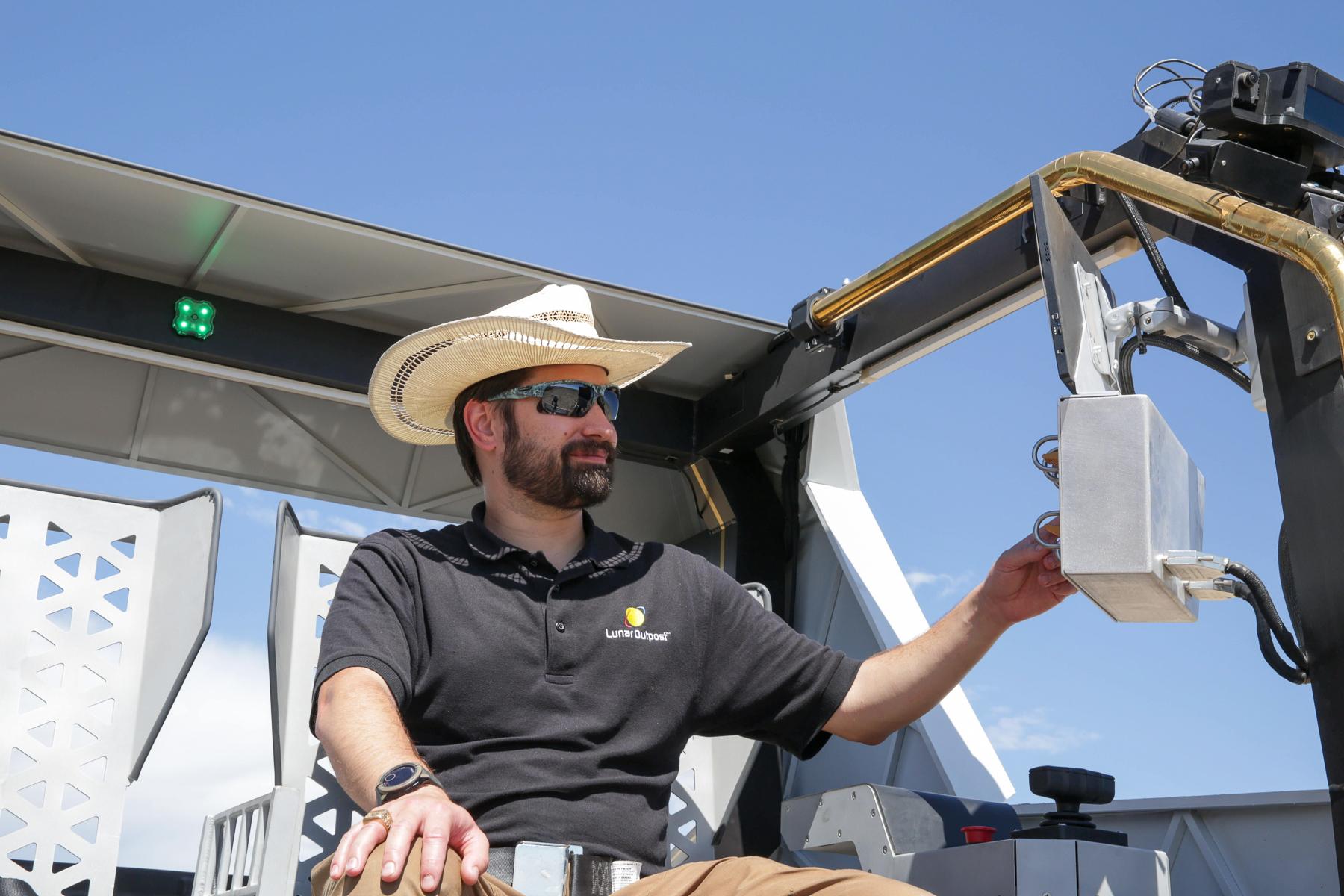 A man wearing sunglasses and a cowboy hat flips switches on a control panel while sitting in a moon rover prototype cockpit.