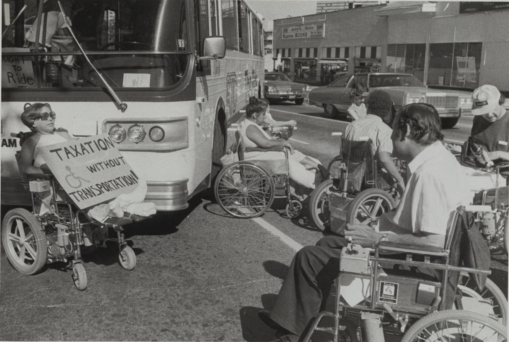 A group of protesters who use wheelchairs stop traffic to raise awareness of the lack of accessibility regarding the Regional Transportation District (RTD) buses. A sign on the left reads: "Taxation without transportation!" with the handicapped symbol drawn on the poster.