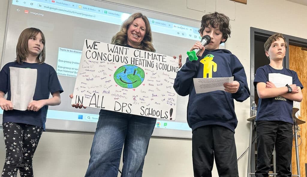 A young boy holds a microphone to his mouth as two other students and a teacher look on. The teacher holds a poster board with a drawing of Earth that says &quot;We want climate conscious heating &amp; cooling in all DPS schools.&quot;