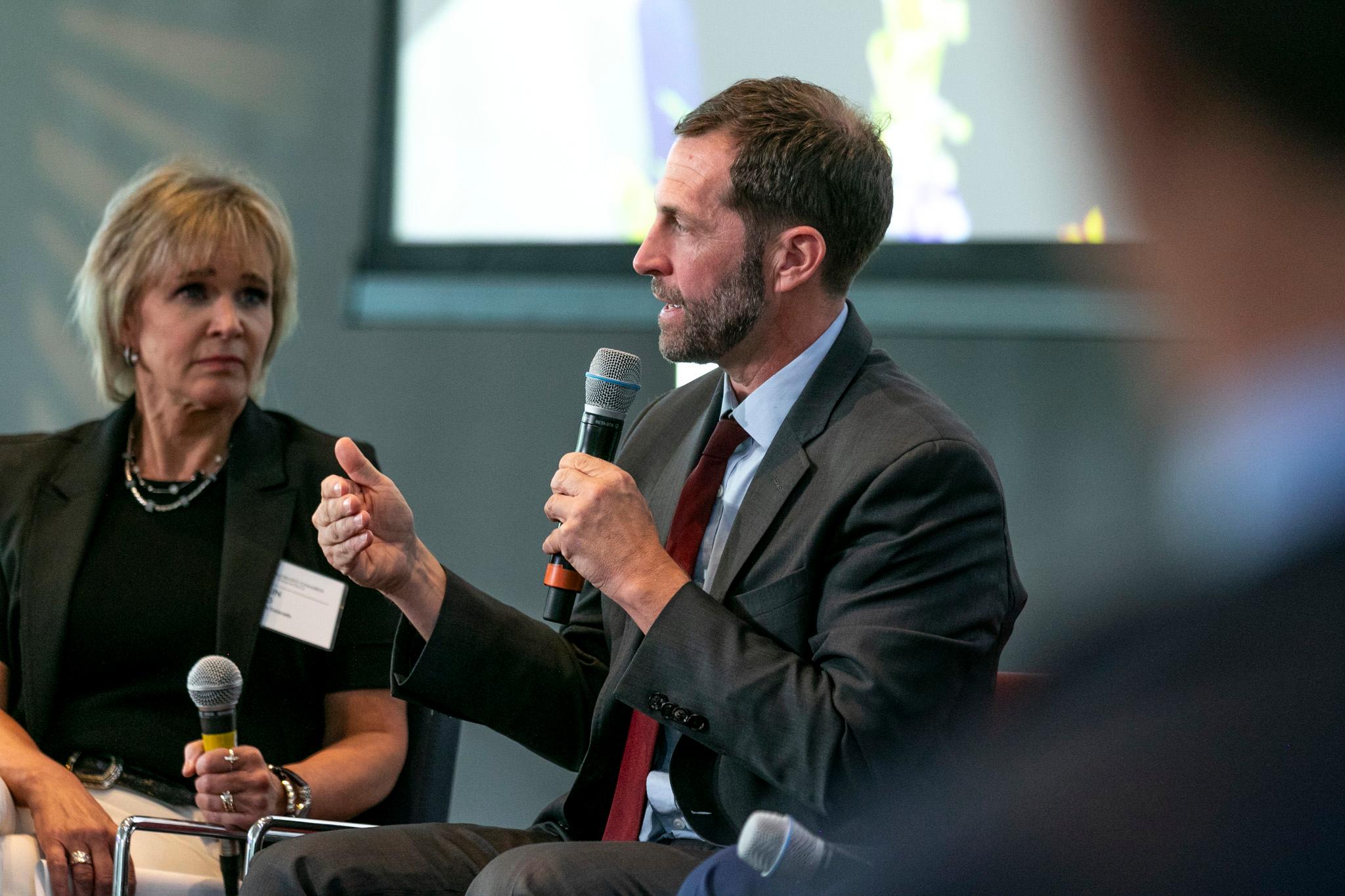 A man and a woman are sitting in front of a microphone, with the man speaking into it.
