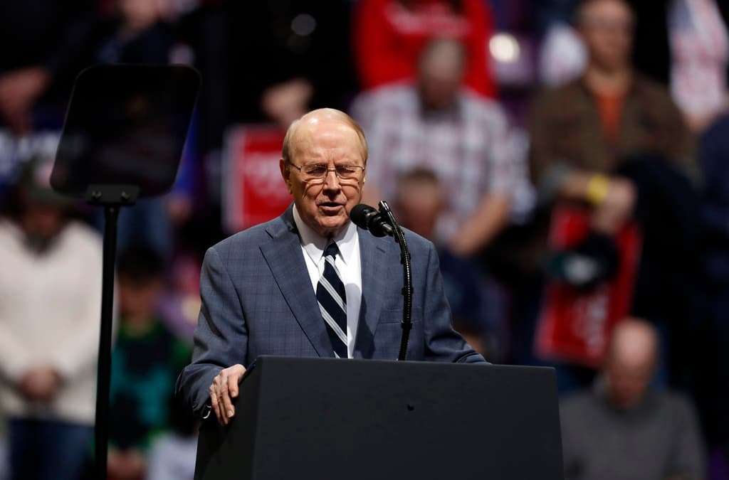 A man in a blue suit stands at a lectern with his eyes closed.