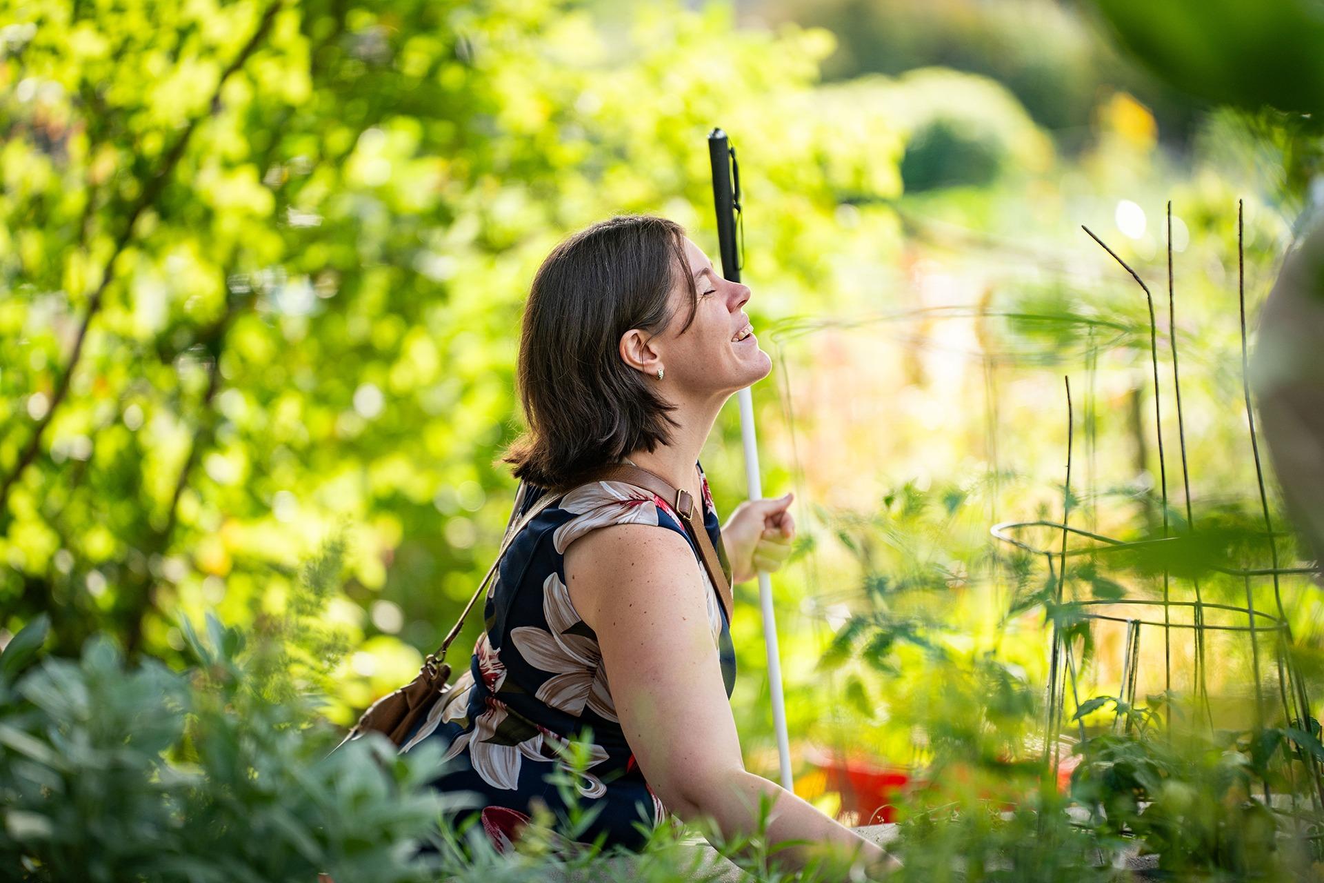 A woman holding a cane sits in a garden we see her side profile