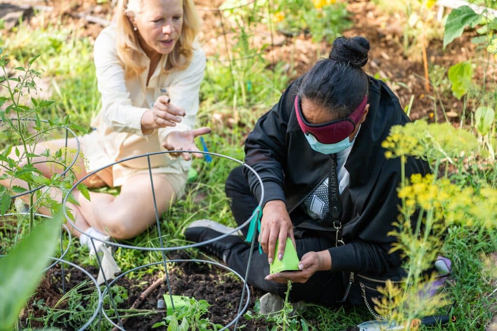 Two women sit on the ground in a garden. One has a blindfold over their eyes