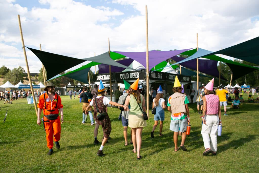 A group of 7 people in pointy elfish hats frolic in a field, walking towards the &quot;cold beer&quot; and &quot;cocktail&quot; tent near the main stage.