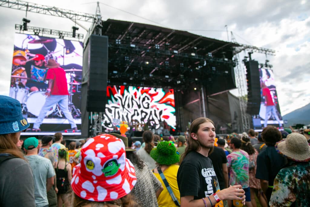 A stage with a big, &quot;King Stingray&quot; backdrop is surrounded by hundreds of fans. In the foreground, a man faces away from the stage, staring into the distance, as everyone around him looks towards the music.