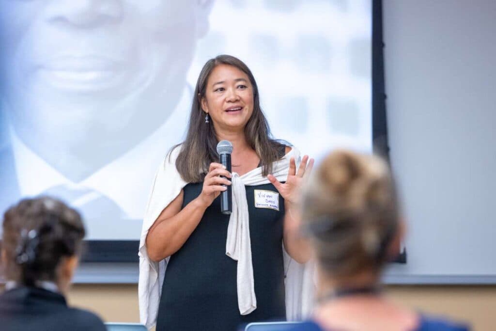 Woman in a navy dress with a white cardigan over her shoulder with a mic in her hands at the front of a classroom