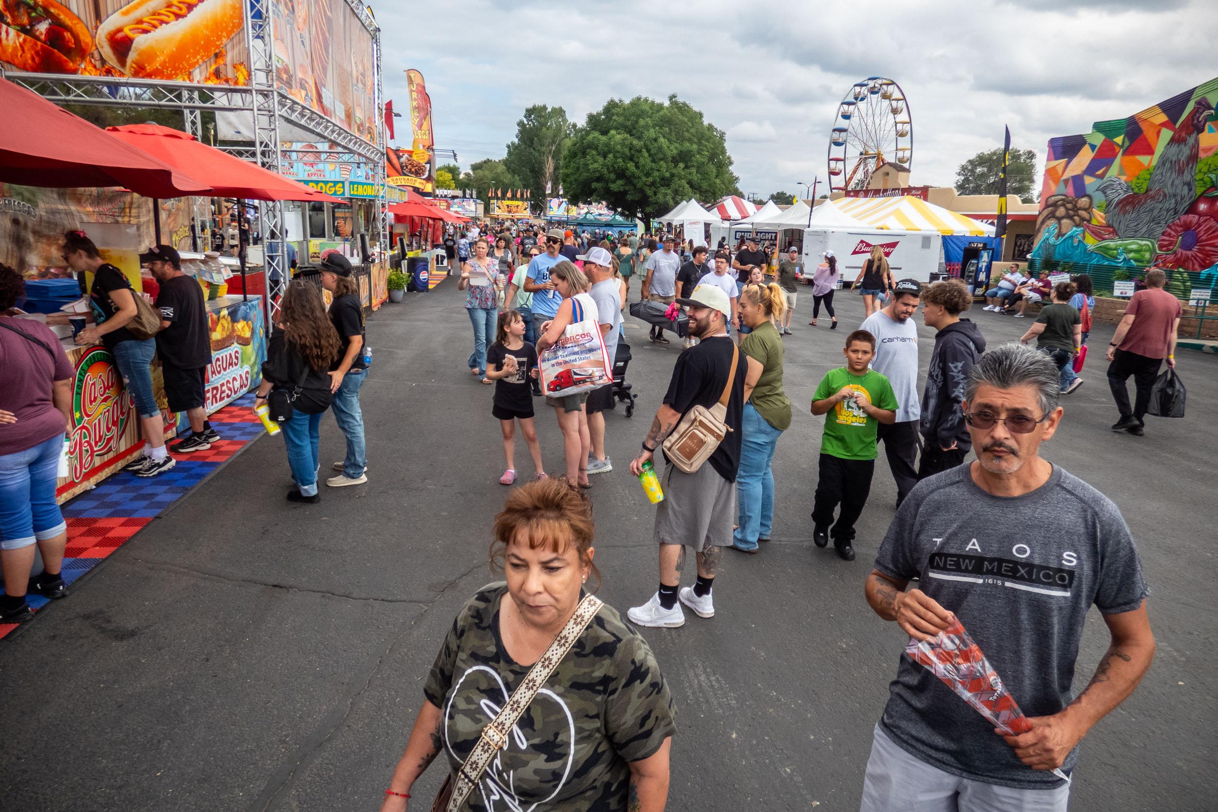 crowds walk between food booths with a ferris wheel in the background