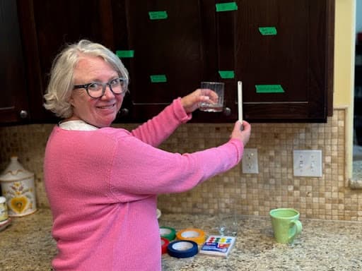 A woman wearing a pink sweater looks toward the camera. Her arms are outstretched and she's holding a glass and a ruler. She is in a kitchen with dark cabinets with bits of green tape on the cabinets.