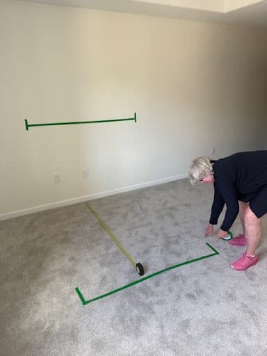 Photos shows an empty room with green tape on the wall and carpet. A woman wearing black is bending down on the right side of the image to put more tape on the carpet.