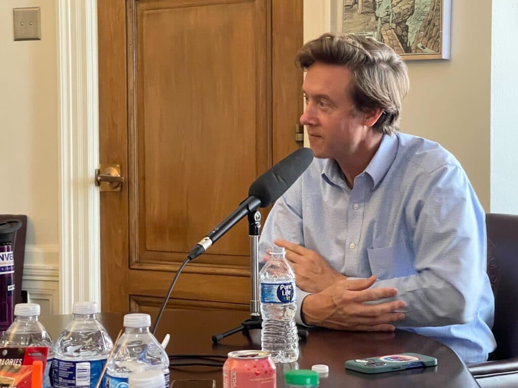 Photos shows Denver Mayor Mike Johnston seated in front of a microphone on a table. There is a wooden door in the background. Johnston is looking to the left.