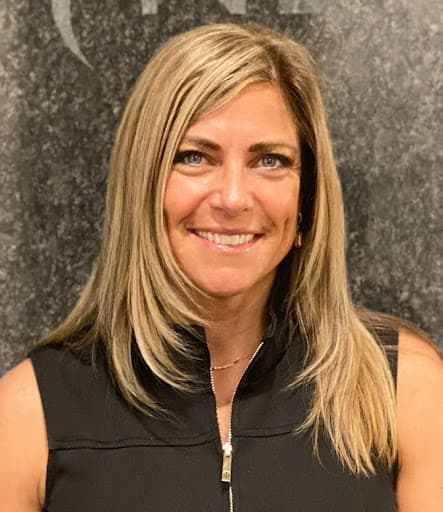 Photo is a head and shoulders image of a woman with blonde hair smiling at the camera. She is in front of a gray background.