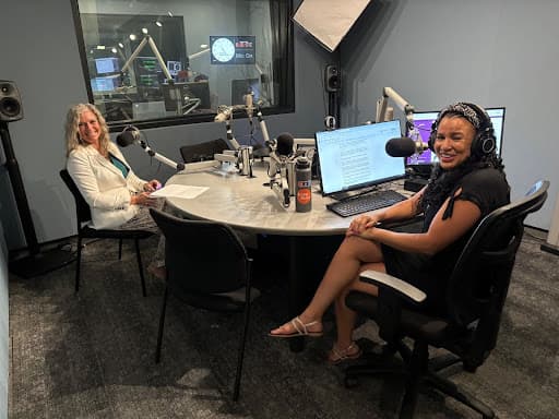 Image shows a CPR News studio. Seated at the left is a woman with blonde hair in a white suit. On the right is Colorado Matters co-host Chandra Thomas Whitfield in a chair wearing black. Both are looking toward the camera. There's a computer screen next to Chandra.